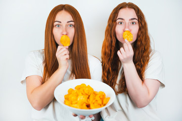 two redheaded young women holding crispy potatoe chips near their mouths standing on isolated white backgroung, junk food concept