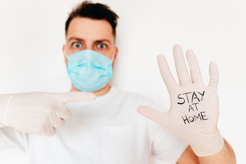 A young european man in blue mask calls for quarantine with sign on protective gloves. Stay at home. Corona virus protection. Covid-19 prevention