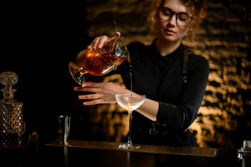 Professional lady barman masterfully pours cocktail from mixing cup into glass.