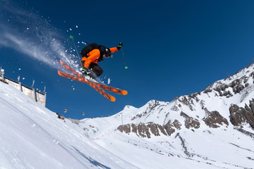 Naklejka na ściany i meble A male athlete skier in an orange trigger makes a jump jump with a grab with flying snow powder against the background of Mount Elbrus in the North Caucasus. Winter Extreme Sports Concept