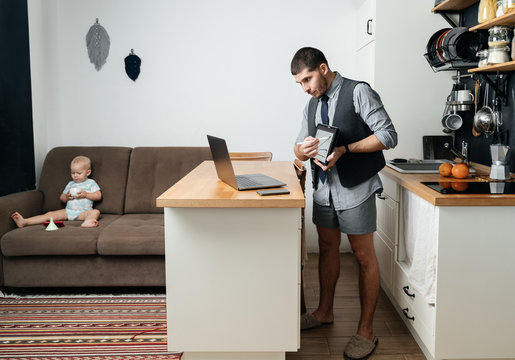 Remote work at home. A man at a laptop with a tablet works online in home shorts and a shirt with a tie against the background of the kitchen interior with a child.