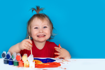 A cute child with a brush in his hands laughs merrily. The girl draws with colored paints on a white sheet, the foreground is blurred. Happy childhood. Copy space