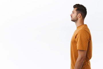 Profile studio shot attractive young modern hipster guy in brown stylish t-shirt standing in line to buy coffee, waiting someone as looking left casually, no emotions, standing white background