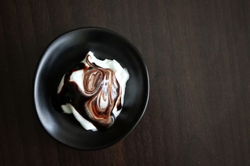 Closeup of vanilla ice cream with chocolate topping in black bowl on dark wooden table. Macro, flat lay, top view.