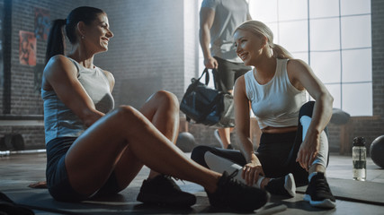 Two Beautiful Fit Athletic Girls Sit on a Floor of Industrial Loft Gym. They're Happy with their Training Program and Successfully Give a High Five. Strong Masculine Man Walks in the Background.