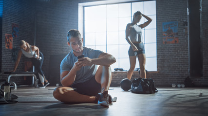 Handsome and Masculine Athletic Young Man is Using a Smartphone while Sitting on a Floor in a Loft Gym. He's Typing a Message and Thinking. He has Sweat on Forehead. Women Exercise in the Background. 