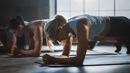 Fotobehang Sportschool Strong Masculine Man and Two Fit Atletic Women Hold a Plank Position in Order to Exercise Their Core Strength. Young Blond Girl is in the Foreground. They Workout in a Loft Gym.  © Gorodenkoff