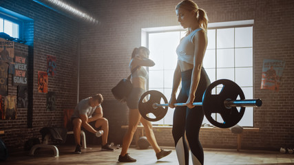 Fototapeta na wymiar Beautiful Fit Athletic Blond Female is Lifting a Barbell from the Ground and Doing Back Strengthening Exercise as Her Daily Fitness Training Routine. She's Working Out in Loft Gym with Sporty People.