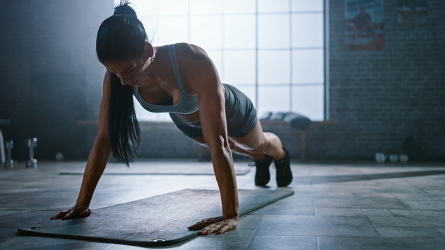 Strong and Fit Athletic Busty Woman in Sport Top and Shorts is Doing Push Up Exercises in a Loft Style Industrial Gym with Motivational Posters. It's Part of Her Cross Fitness Training Workout. - Powered by Adobe