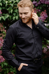 Red-haired elegant smiling man with a beard in a black suit posing in a lilac garden
