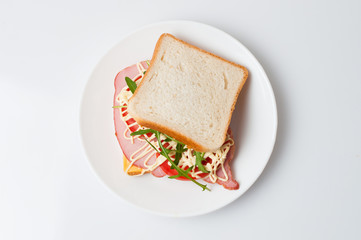 Tasty sandwich with ham or balyk, tomato, arugula, cheese, mayonnaise and white bread for toast on a plate on a white background view from the top flat lay, copy space