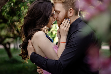 Fototapeta premium Beautiful couple in love cuddling in a blooming lilac garden. A woman in a purple sleeveless dress with a delicate necklace around her neck. Red-haired man in a black suit kisses and hugs a girl