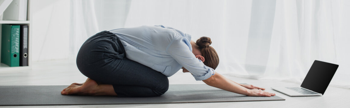 panoramic shot of young businesswoman in Child Pose having online yoga classes on laptop in office