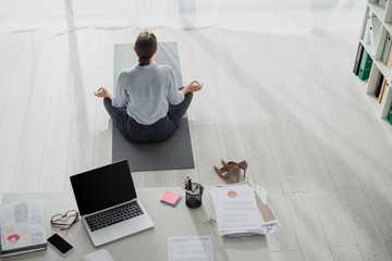 Back view of businesswoman practicing yoga in lotus position with gyan mudra on mat in office with laptop