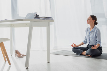 Young businesswoman practicing yoga in lotus position with gyan mudra on mat in office