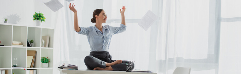 panoramic shot of happy businesswoman sitting in lotus pose on table with falling business papers...