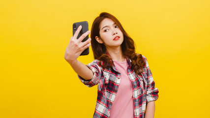 Smiling adorable Asian female making selfie photo on smartphone with positive expression in casual clothing and looking at camera over yellow background. Happy adorable glad woman rejoices success.