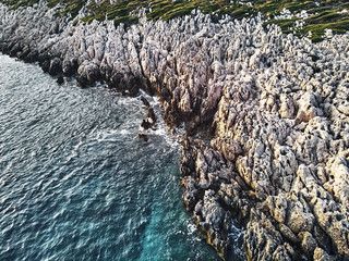 top drone view of a stony coast clashing into the blue ocean sea. Structure and Pattern in artistic way of the sharp rocks in the water. Aerial top shot on warm summer waves.