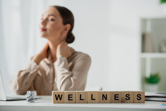 selective focus of businesswoman resting at workplace with laptop and alphabet cubes with wellness word