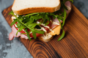 Very tasty big sandwich with ham or balyk, arugula, cheese, tomatoes and mayonnaise on a cutting board in rustic style on a gray background view from the top, copy space