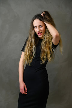 Vertical photo of a slender young Caucasian girl model with long curly hair in a black dress on a gray background. Made in the studio.