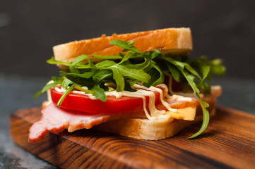 Very tasty big sandwich with ham or balyk, arugula, cheese, tomatoes and mayonnaise on a cutting board in rustic style on a gray background view from the side, copy space