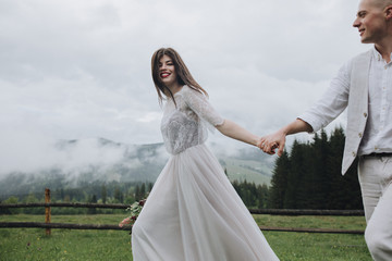 Fototapeta na wymiar Spring wedding in the mountains. A guy in a shirt and vest and a girl in a white dress walk along a wooden fence along a green meadow against the backdrop of mountains and forests