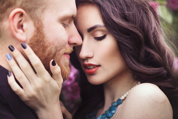 Beautiful couple in love cuddling in a blooming lilac garden. A woman in a purple sleeveless dress with a delicate necklace around her neck. Red-haired man in a black suit kisses and hugs a girl