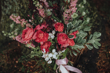 Wedding floristry. A bouquet of red and white flowers with greenery and a pink ribbon stands on the ground near a tree covered with moss.
