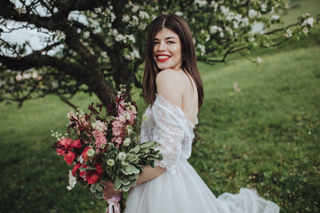 Spring wedding in the mountains. A young girl in a white dress stands near a blossoming tree in the mountains and holds in her hands a bouquet of red flowers with a ribbon