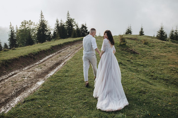 Spring wedding in the mountains. A guy in a shirt and vest and a girl in a white dress walk along a mountain road at sunset among the clouds