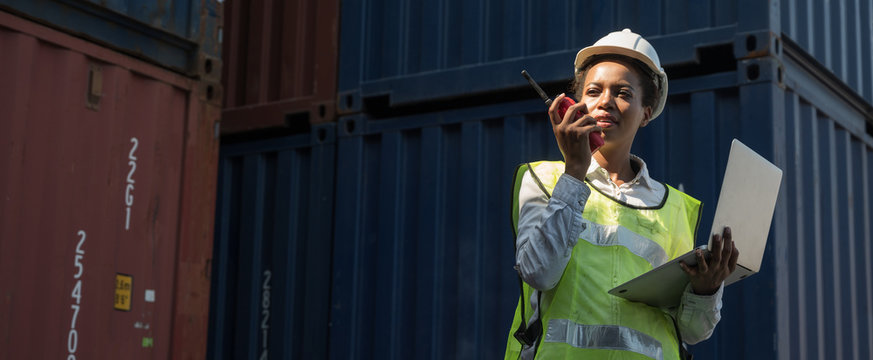 Black foreman woman worker working checking at Container cargo harbor holding laptop and radio to loading containers. African dock female staff business Logistics import export shipping concept.