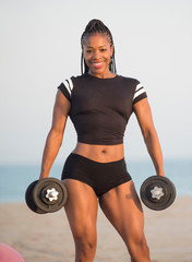 Beautiful African American Black female fitness model on the beach wearing black sportswear holding a pair of silver dumbbells looking at the camera 