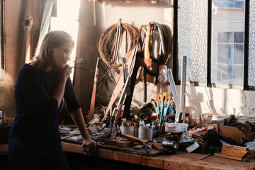 A beautiful young woman taking off her glasses in the sunlight stands in the workshop thinking about a new product