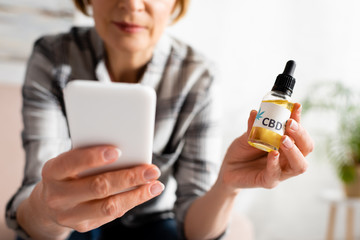 cropped view of mature woman holding bottle with cbd lettering and using smartphone