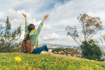 Freedom and tourism concept. Rear view of a girl sitting on grass with raised arms looking at sea and mountain panorama.