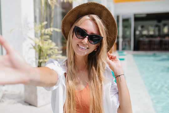 Smiling gorgeous girl with long hair chilling at resort and taking picture of herself. Romantic caucasian female model making selfie near pool in hot day.