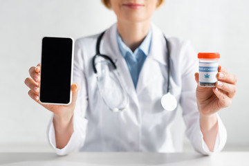 cropped view of mature doctor in white coat holding smartphone with blank screen and bottle with medical cannabis lettering