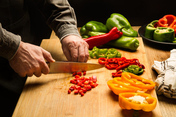 Manos de hombre con un cuchillo cortando un pimientos rojo, amarillo y verde sobre una tabla de madera rústica. Vista superior