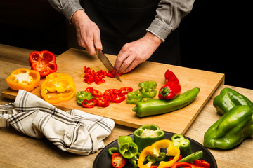 Manos de hombre con un cuchillo cortando un pimientos rojo, amarillo y verde sobre una tabla de madera rústica. Vista superior