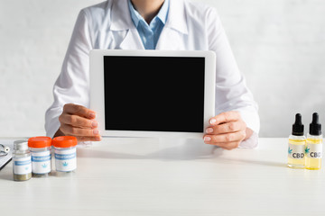 cropped view of doctor holding digital tablet with blank screen near bottles with cbd and medical cannabis lettering in clinic