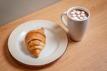 A mug of cocoa with marshmallows on a wooden table next to a croissant.