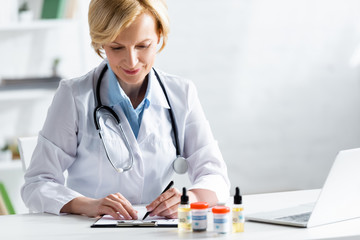 selective focus of cheerful and mature doctor writing prescription near bottles with medical cannabis and cbd