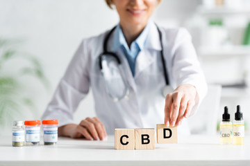 cropped view of happy doctor in white coat touching cubes with cbd lettering near drugs