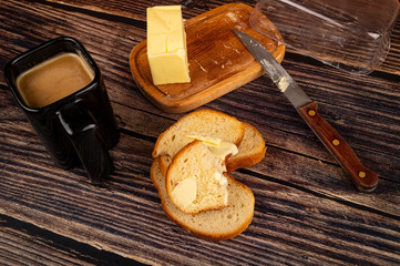 Fresh wheat toast with butter, a ceramic mug of coffee with milk and a piece of butter in a wooden butter dish on a wooden background. Close up.