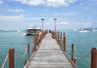 Naklejka premium A long wooden pier at the ferry from Koh Samui to Phangan Island in Thailand. A ferry-boat departs from the pier, several yachts float on the horizon on a clear sunny day.