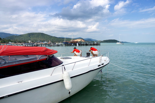 Two men in white and red uniform clean a white yacht moored at the dock of Koh Samui in Thailand. Beautiful view of the wooden pier and mountains covered in greenery. - Powered by Adobe