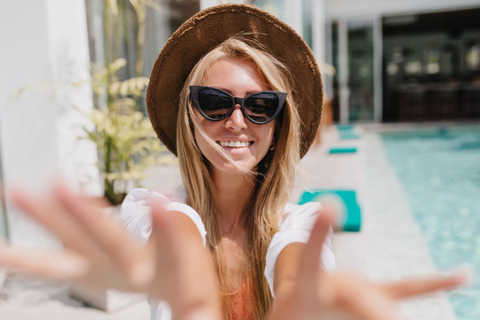 Ecstatic white woman in hat making selfie at resort with wonderful smile. Beautiful european female tourist having fun near pool.