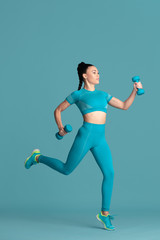 In jump. Beautiful young female athlete practicing in studio, monochrome blue portrait. Sportive fit brunette model with weights. Body building, healthy lifestyle, beauty and action concept.