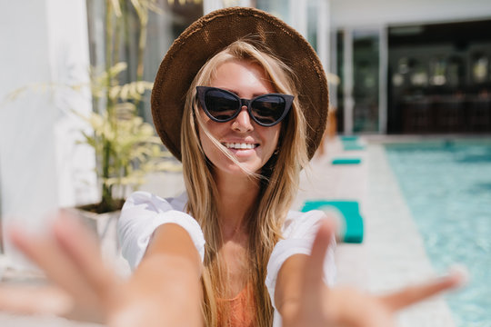 Attractive european girl in black sunglasses smiling while making selfie near swimming pool. Outdoor photo of good-looking female tourist chilling at resort in hot morning.